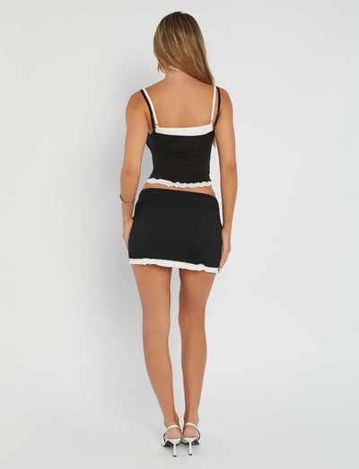 A woman is standing with her back to the camera, wearing the fashionable ADAIR TOP in black and white from TIGERMIST, paired with a matching mini skirt. She completes her chic look with white high-heeled shoes against a plain white background.