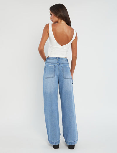 A person with long dark hair is facing away from the camera, wearing the chic GWYN TOP - WHITE by TIGERMIST, featuring a deep V-cut back, paired with high-waisted, light-wash wide-leg jeans. Styled in trendy Tiger Mist fashion, they stand against a plain white background.