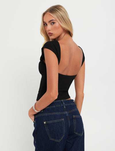 A woman with long blonde hair, styled flawlessly, stands facing slightly away from the camera, looking over her shoulder. She is wearing a chic RYLAN TOP - BLACK with a low back and blue jeans from TIGERMIST. The background is plain white.