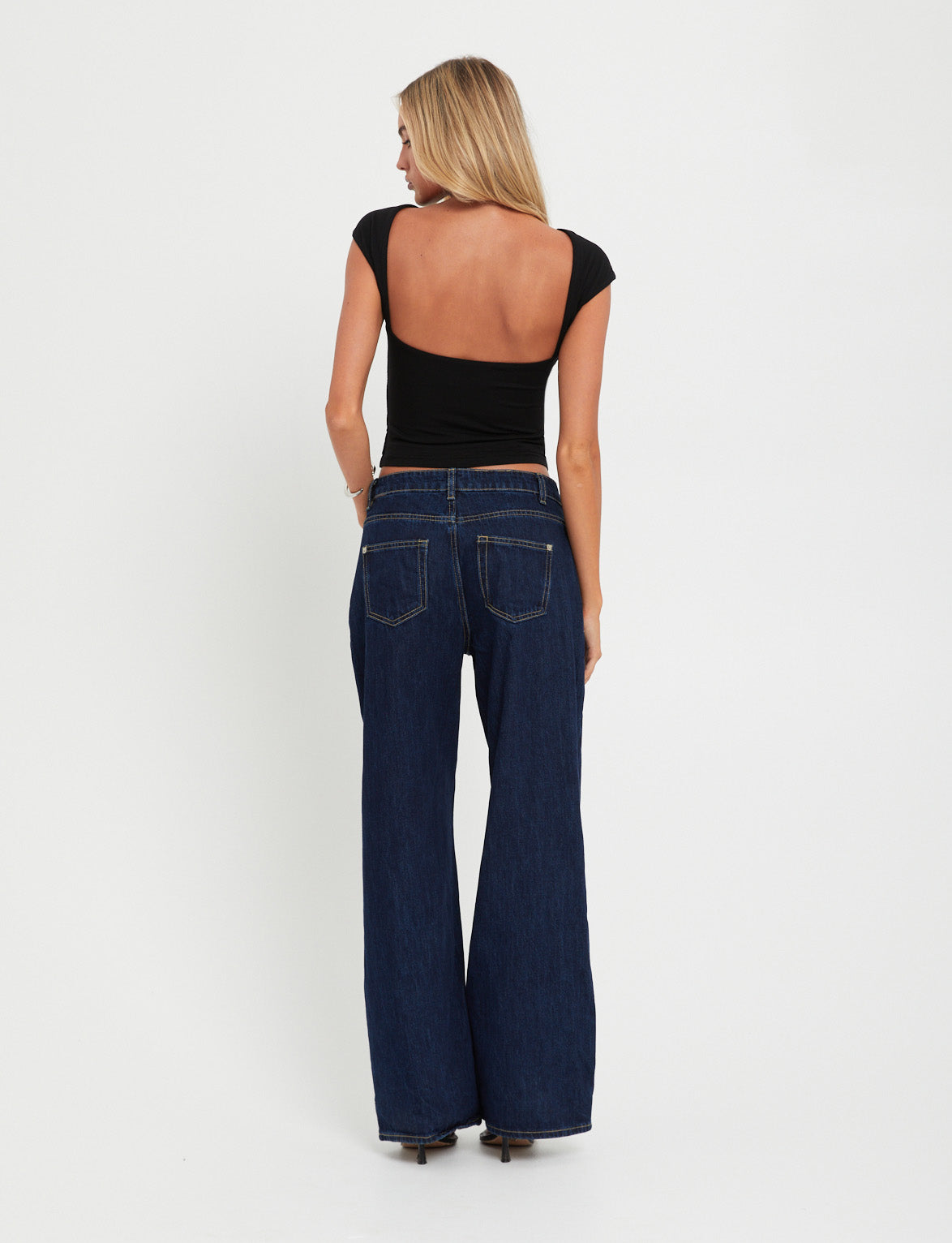 A woman stands facing away, showcasing her back and jeans. She has long blonde hair and is wearing the RYLAN TOP - BLACK from TIGERMIST, a fitted black top with a deep scoop back, paired with dark blue wide-leg jeans. The background is plain white.