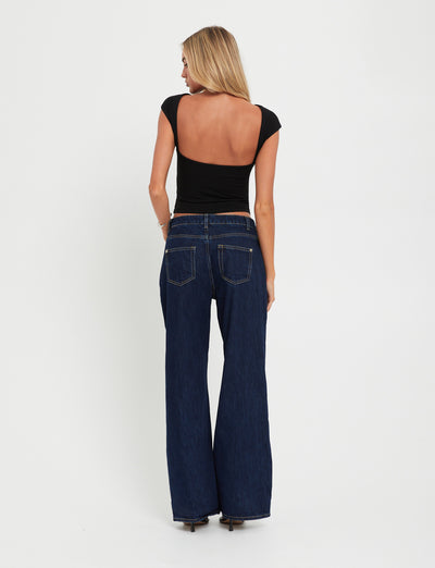 A woman stands facing away, showcasing her back and jeans. She has long blonde hair and is wearing the RYLAN TOP - BLACK from TIGERMIST, a fitted black top with a deep scoop back, paired with dark blue wide-leg jeans. The background is plain white.