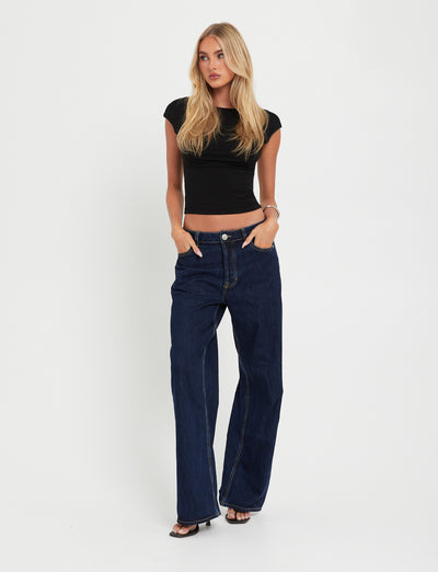 A person with long blonde hair stands against a plain white background, wearing the fitted RYLAN TOP - BLACK and high-waisted dark blue wide-leg jeans from TigerMist. They have their hands in their pockets and are wearing open-toed black shoes.
