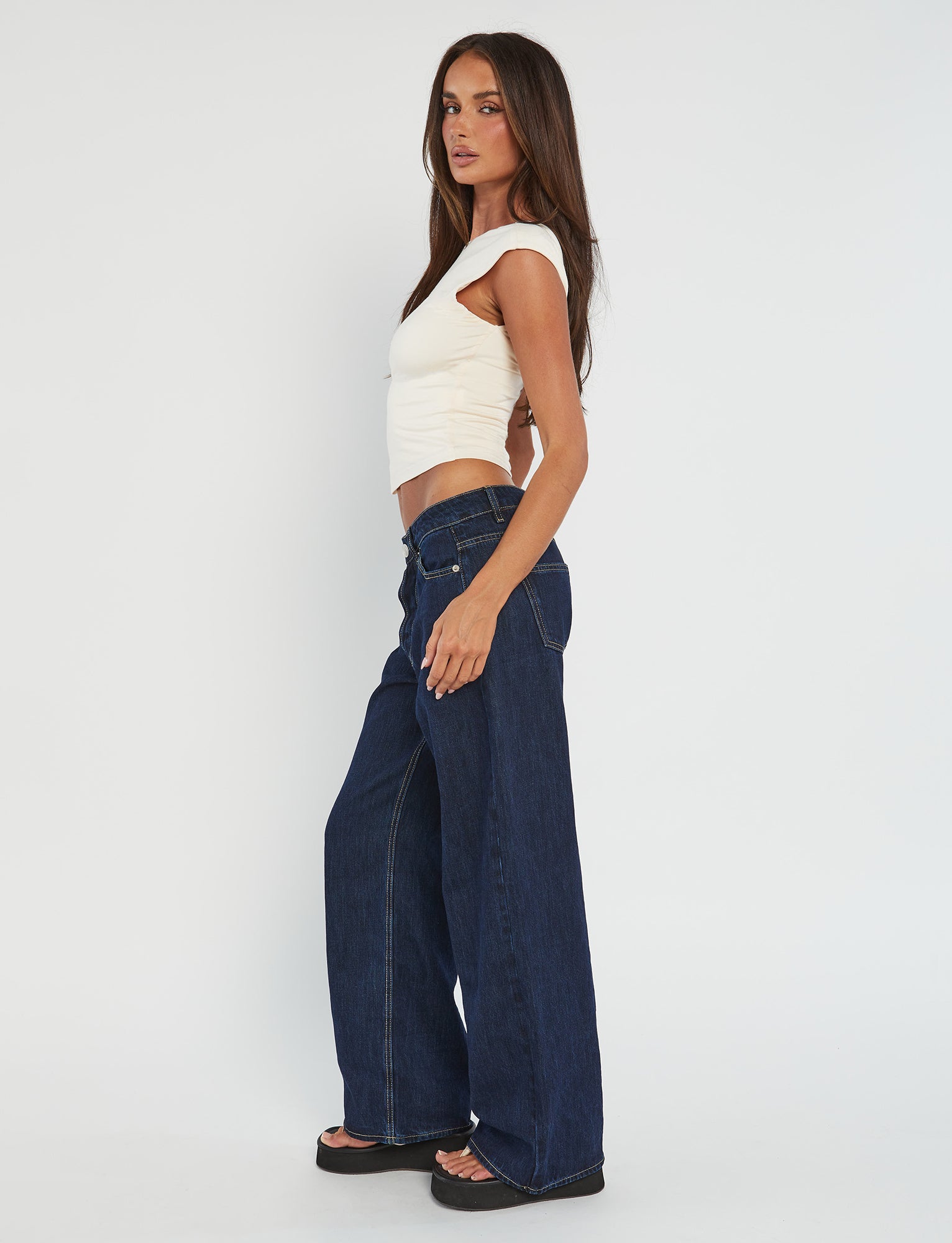 A woman with long brown hair stands in profile, looking at the camera. She is wearing the RYLAN TOP in Beige Sand, a fitted crop top from Tiger Mist, and high-waisted, wide-leg dark blue jeans paired with black platform sandals. The background is plain white.