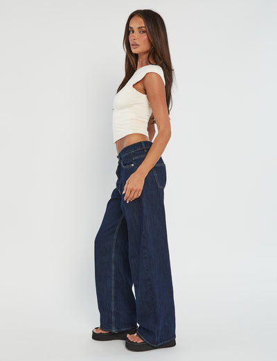A woman with long brown hair stands in profile, looking at the camera. She is wearing the RYLAN TOP in Beige Sand, a fitted crop top from Tiger Mist, and high-waisted, wide-leg dark blue jeans paired with black platform sandals. The background is plain white.