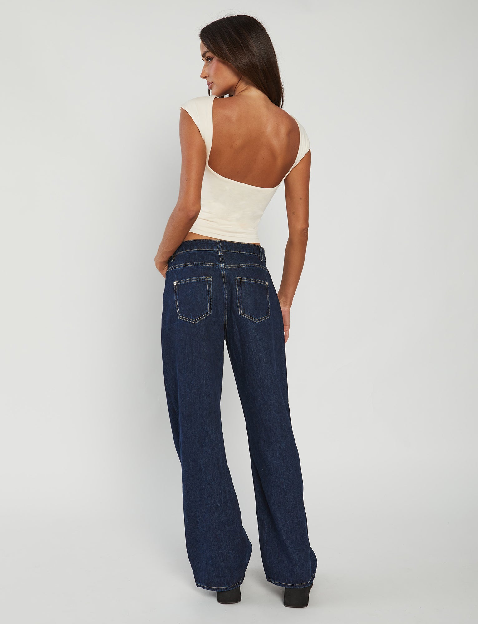 A person with long dark hair is seen from the back, wearing the RYLAN TOP - BEIGE : SAND by TIGERMIST and wide-leg dark blue jeans with back pockets and yellow stitching. The background is a plain light grey.