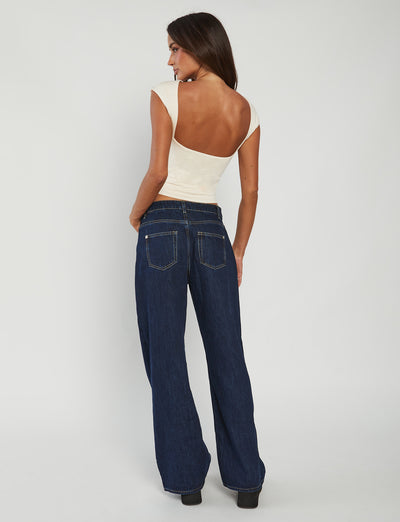 A person with long dark hair is seen from the back, wearing the RYLAN TOP - BEIGE : SAND by TIGERMIST and wide-leg dark blue jeans with back pockets and yellow stitching. The background is a plain light grey.