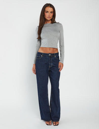 A woman with long, dark hair stands against a white background. She is wearing the fitted QUINNELL TOP in grey from TIGERMIST and high-waisted dark blue wide-leg jeans. She appears to be looking directly at the camera, with her arms relaxed by her sides.