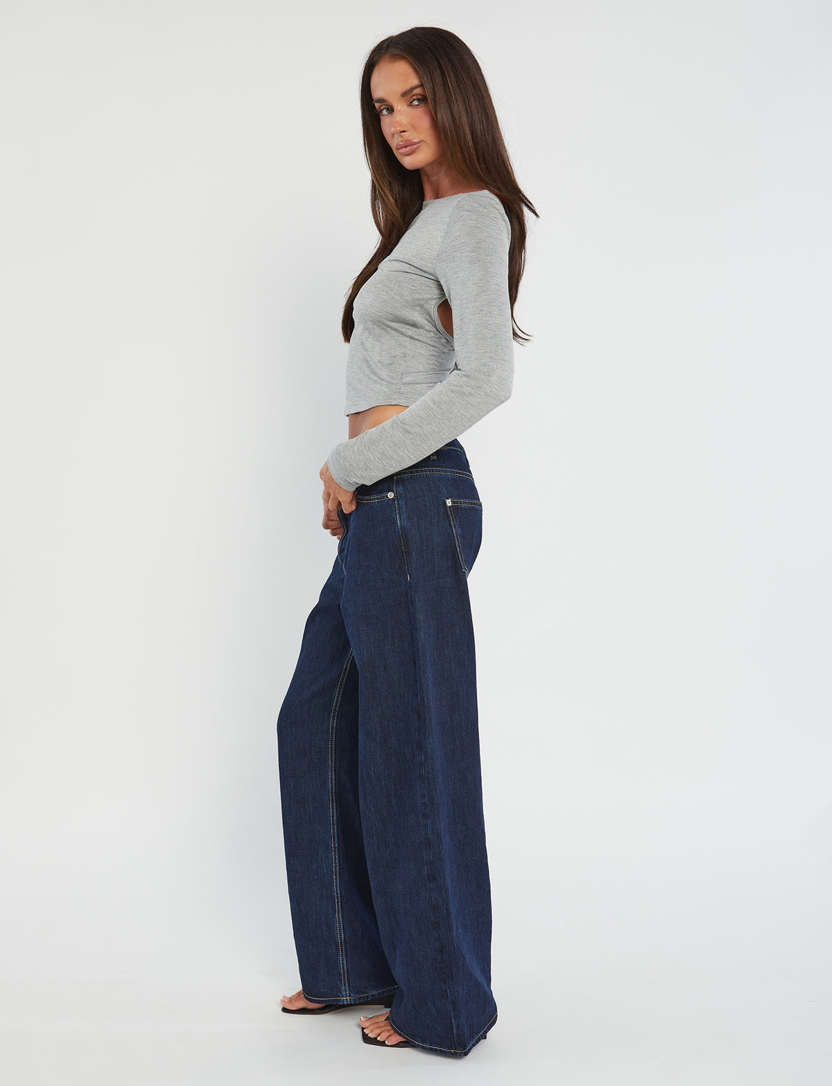 A woman stands sideways against a plain white background, wearing the QUINNELL TOP - GREY and wide-leg blue jeans from TIGERMIST. She is also wearing black heels and has straight, long brown hair. Her gaze is directed towards the camera while her arms hang naturally by her sides.