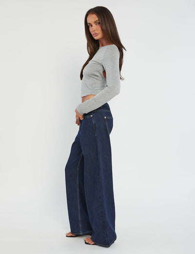 A woman stands sideways against a plain white background, wearing the QUINNELL TOP - GREY and wide-leg blue jeans from TIGERMIST. She is also wearing black heels and has straight, long brown hair. Her gaze is directed towards the camera while her arms hang naturally by her sides.