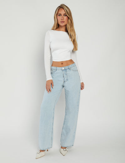 A woman stands against a plain white background, wearing the QUINNELL TOP - WHITE from TIGERMIST, a long-sleeved, fitted white crop top. She pairs it with light blue high-waisted wide-leg jeans and is styled with long, wavy blonde hair. She completes her look with white pointed-toe shoes and poses gracefully with her left hand resting on her hip.