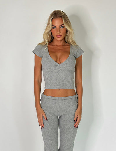 A woman with long blonde hair is standing against a plain white background, wearing the chic Kavat Top in Dove Grey from Tigermist along with matching grey pants. Her hands are relaxed at her sides, and she maintains a neutral expression.