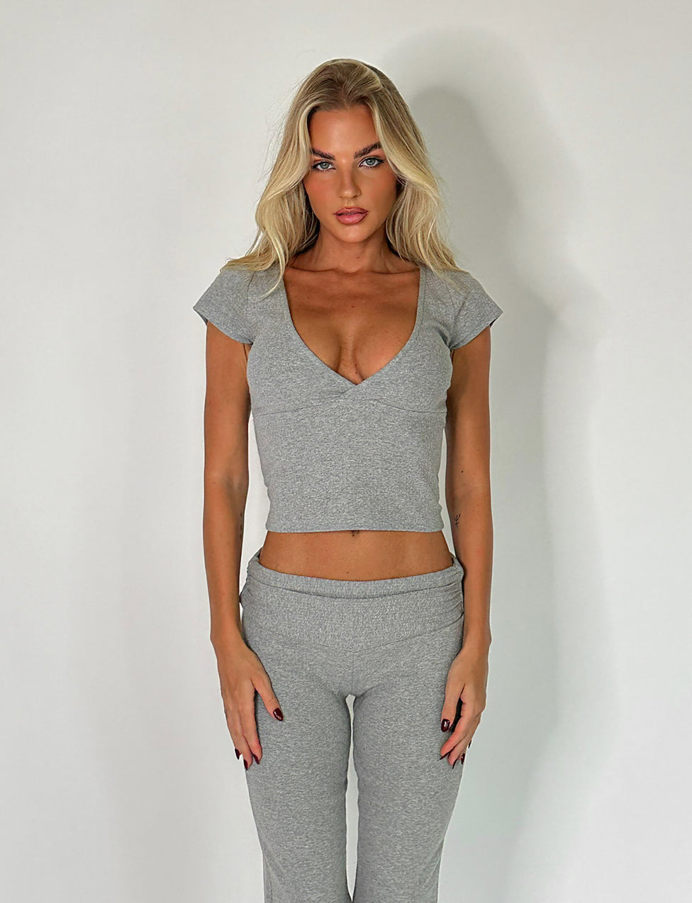 A woman with long blonde hair is standing against a plain white background, wearing the chic Kavat Top in Dove Grey from Tigermist along with matching grey pants. Her hands are relaxed at her sides, and she maintains a neutral expression.