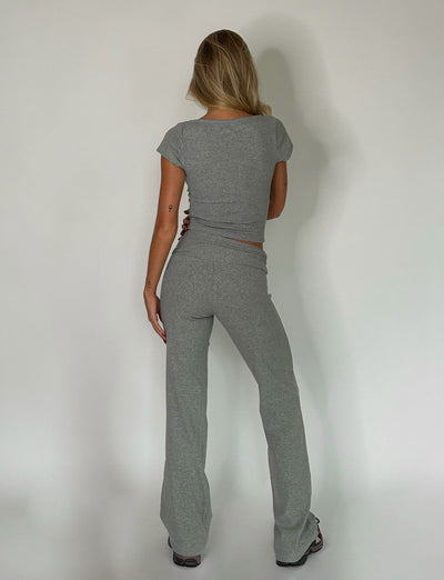 A person with long blonde hair stands facing a white wall, their back to the camera. They are wearing a fitted Dove Grey short-sleeve Kavat Top and matching gray pants from Tiger Mist. The lighting is soft, creating subtle shadows on the wall.