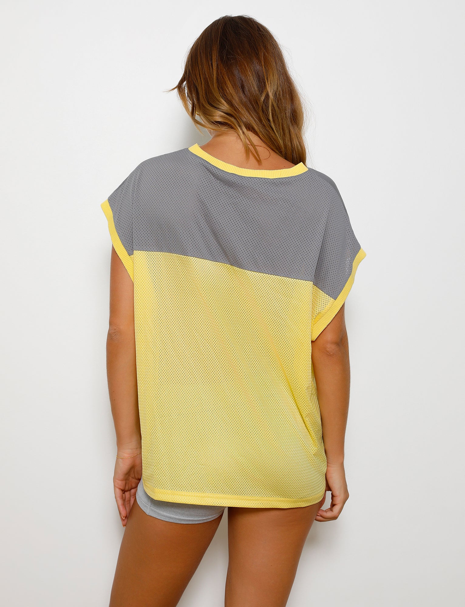 A person with long hair stands facing away from the camera, wearing a loose-fitting, pale yellow Zenith Tank from TigerMist. The tank has short sleeves and the individual pairs it with grey shorts.