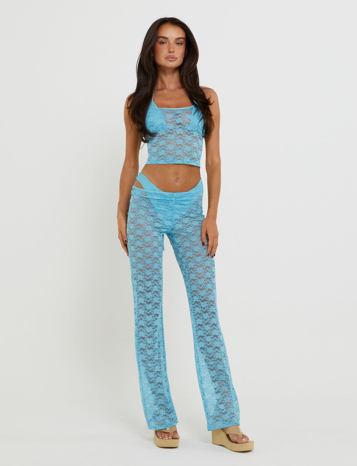 A woman with long dark hair stands against a plain background, wearing the Kaelani Two-Piece Top from TIGERMIST, an ice blue ensemble featuring a bright blue lace halter top and matching flared pants. She pairs the outfit with beige platform sandals and poses with her left hand resting on her hip.