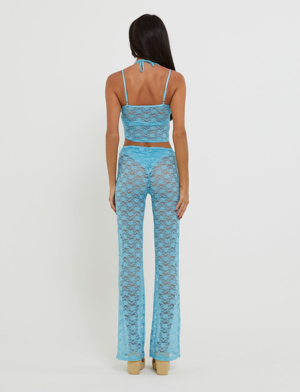 A person with long dark hair is standing with their back to the camera, wearing a fashionable KAELANI TWO-PIECE TOP in ice blue from TIGERMIST. The ensemble features a sleeveless crop top and flared pants. They are poised on light-colored, high-platform shoes against a plain white background.