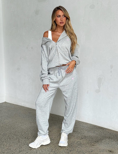 A woman with long blonde hair poses against a plain wall, showcasing her light grey tracksuit from TIGERMIST. She wears a zip-up hoodie that is slightly off her shoulder, revealing a white sports bra underneath and the ALISH PANT in GREY MARLE. White sneakers complete her effortlessly chic look.