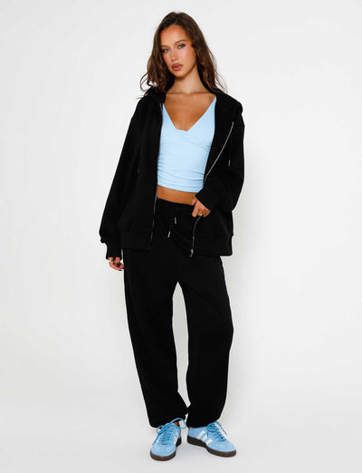 A person with long hair stands against a plain white background, wearing a light blue cropped top from Tiger Mist, the ALISH HOODIE - BLACK by TIGERMIST, and black jogger pants. They are also sporting light blue sneakers. One hand is partially in the hoodie pocket while the other hangs by their side.