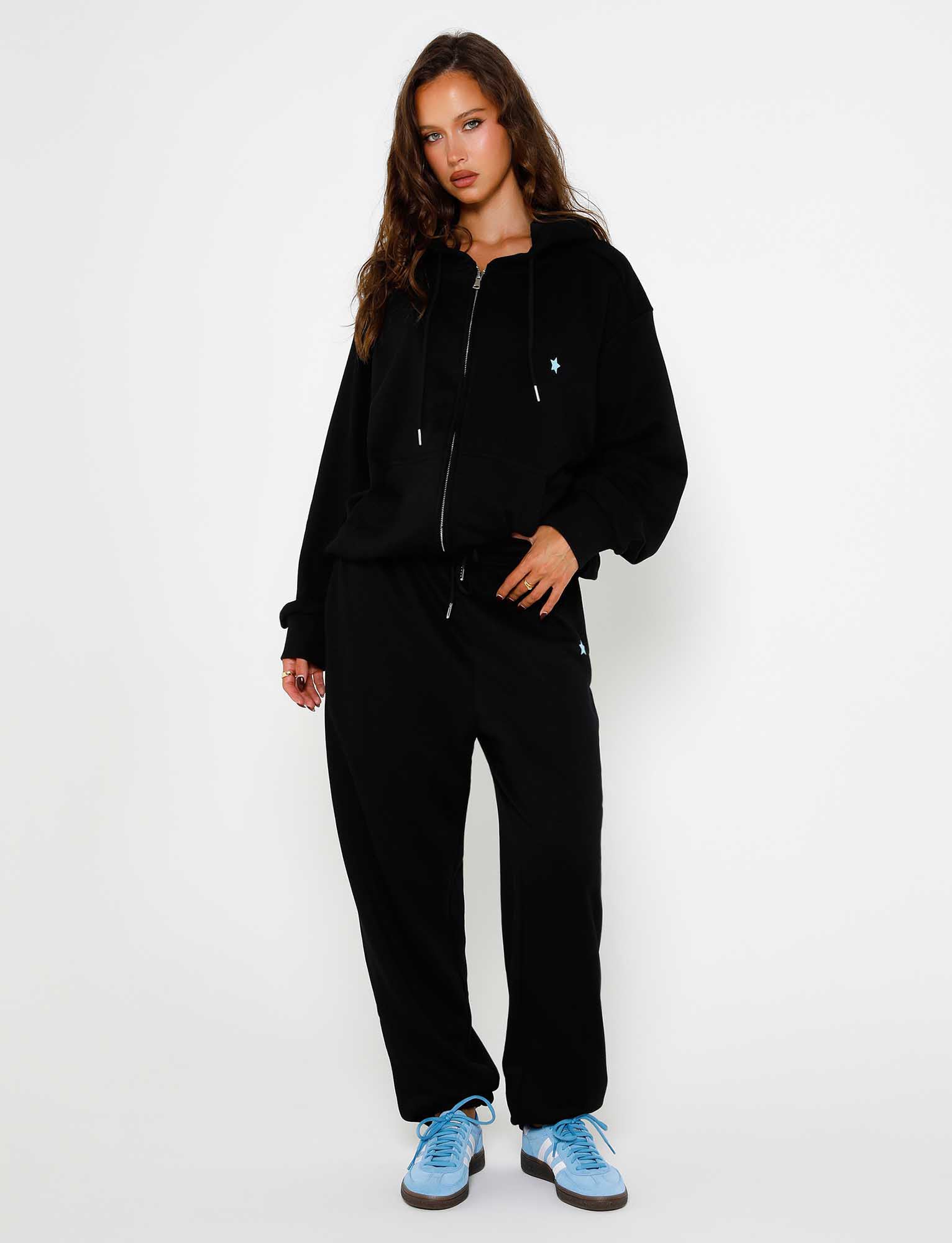 A person with long, wavy hair stands against a plain white background. Wearing the TIGERMIST ALISH HOODIE in black with a small light blue emblem, black jogger pants, and light blue sneakers, one hand rests in their pocket while the other hangs by their side.