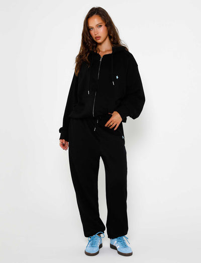 A person with long, wavy hair stands against a plain white background. Wearing the TIGERMIST ALISH HOODIE in black with a small light blue emblem, black jogger pants, and light blue sneakers, one hand rests in their pocket while the other hangs by their side.