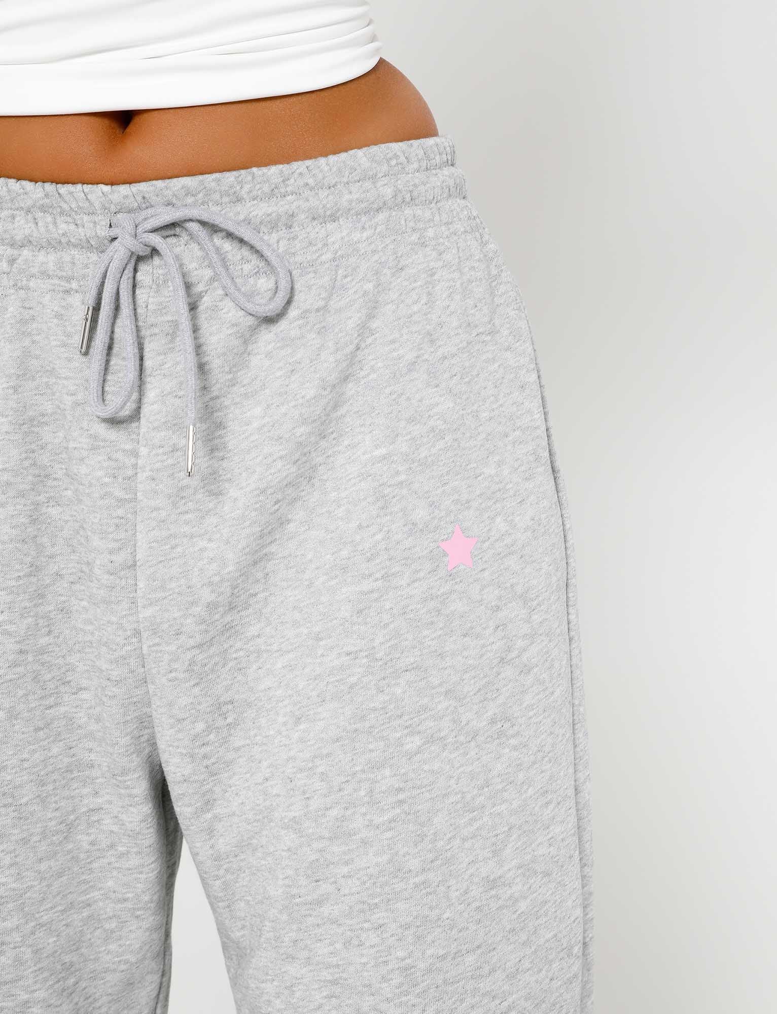 Close-up of a person wearing grey TIGERMIST ALISH PANT - GREY : GREY MARLE sweatpants with a drawstring and a small pink star logo on the upper left thigh area. The person is also wearing a white top, visible just above the waistband of the sweatpants. The background is plain white.