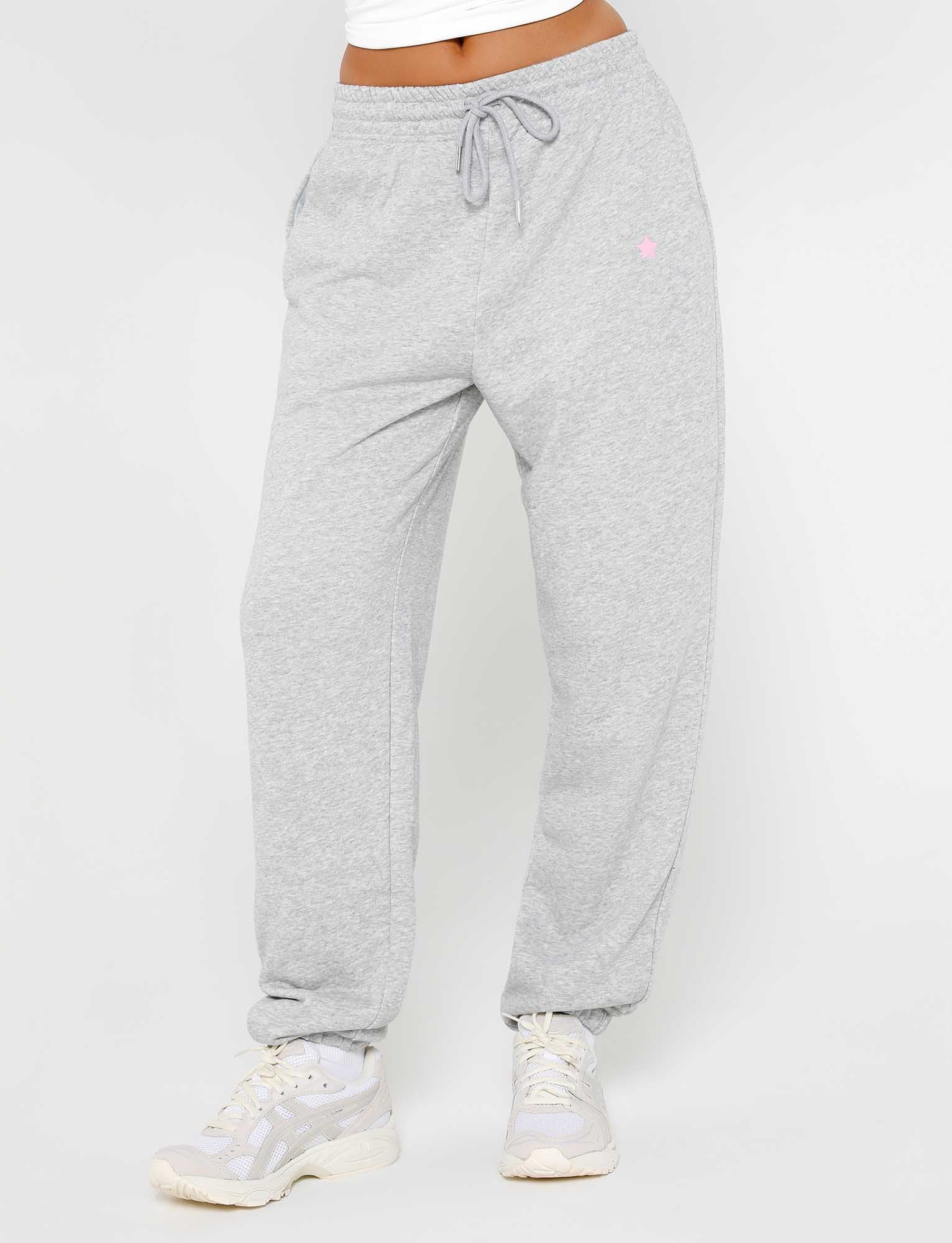 A person wearing the ALISH PANT - GREY : GREY MARLE from TIGERMIST, featuring an elastic waistband and drawstring, along with white and gray athletic shoes. The person's upper body is mostly out of frame. The sweatpants have pockets and a small logo on the left thigh.