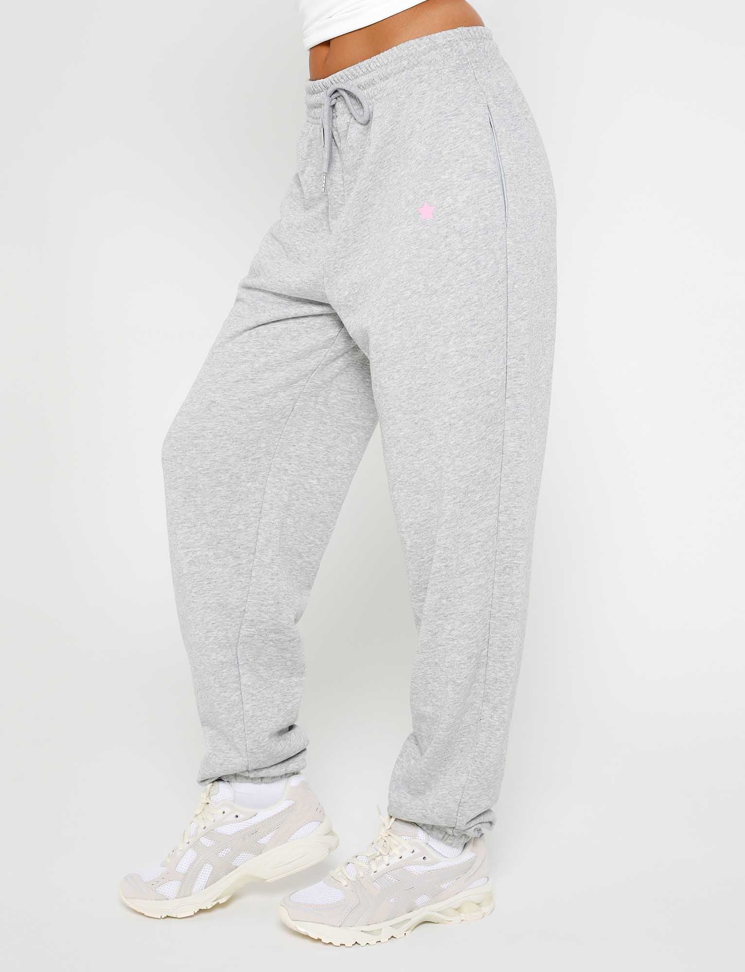 A person is shown from the waist down, wearing the Tiger Mist ALISH PANT in GREY MARLE, featuring an elastic drawstring waistband and complemented by a pair of sneakers. The sweatpants have a small logo on the upper thigh, with a plain white background showcasing the look.