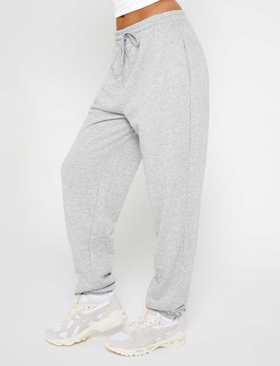 A person is shown from the waist down, wearing the Tiger Mist ALISH PANT in GREY MARLE, featuring an elastic drawstring waistband and complemented by a pair of sneakers. The sweatpants have a small logo on the upper thigh, with a plain white background showcasing the look.