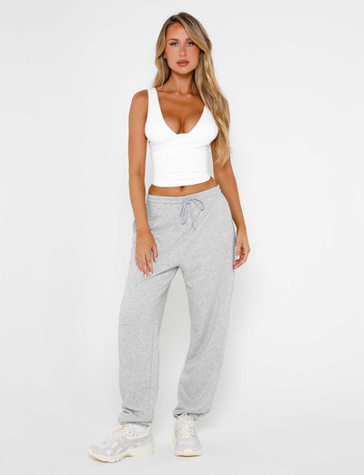 A person with long blonde hair is standing against a plain background, wearing a white sleeveless crop top and the ALISH PANT in Grey Marle from TIGERMIST. They are also wearing white sneakers. Their right hand is resting on their thigh, and they are looking straight ahead.