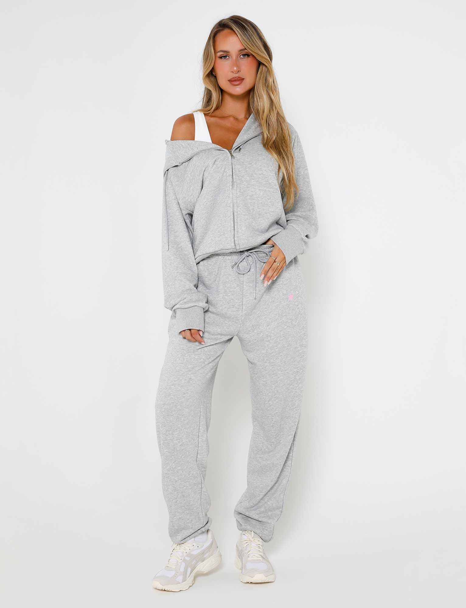 A woman stands against a plain white background, wearing light gray Alish Pant - Grey : Grey Marle from TIGERMIST with a zip-up hoodie and drawstring pants. The hoodie is slightly off one shoulder, revealing a white top underneath. She is also wearing white sneakers. Her hair is long and blonde.