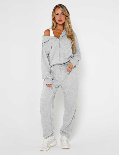 A woman stands against a plain white background, wearing light gray Alish Pant - Grey : Grey Marle from TIGERMIST with a zip-up hoodie and drawstring pants. The hoodie is slightly off one shoulder, revealing a white top underneath. She is also wearing white sneakers. Her hair is long and blonde.