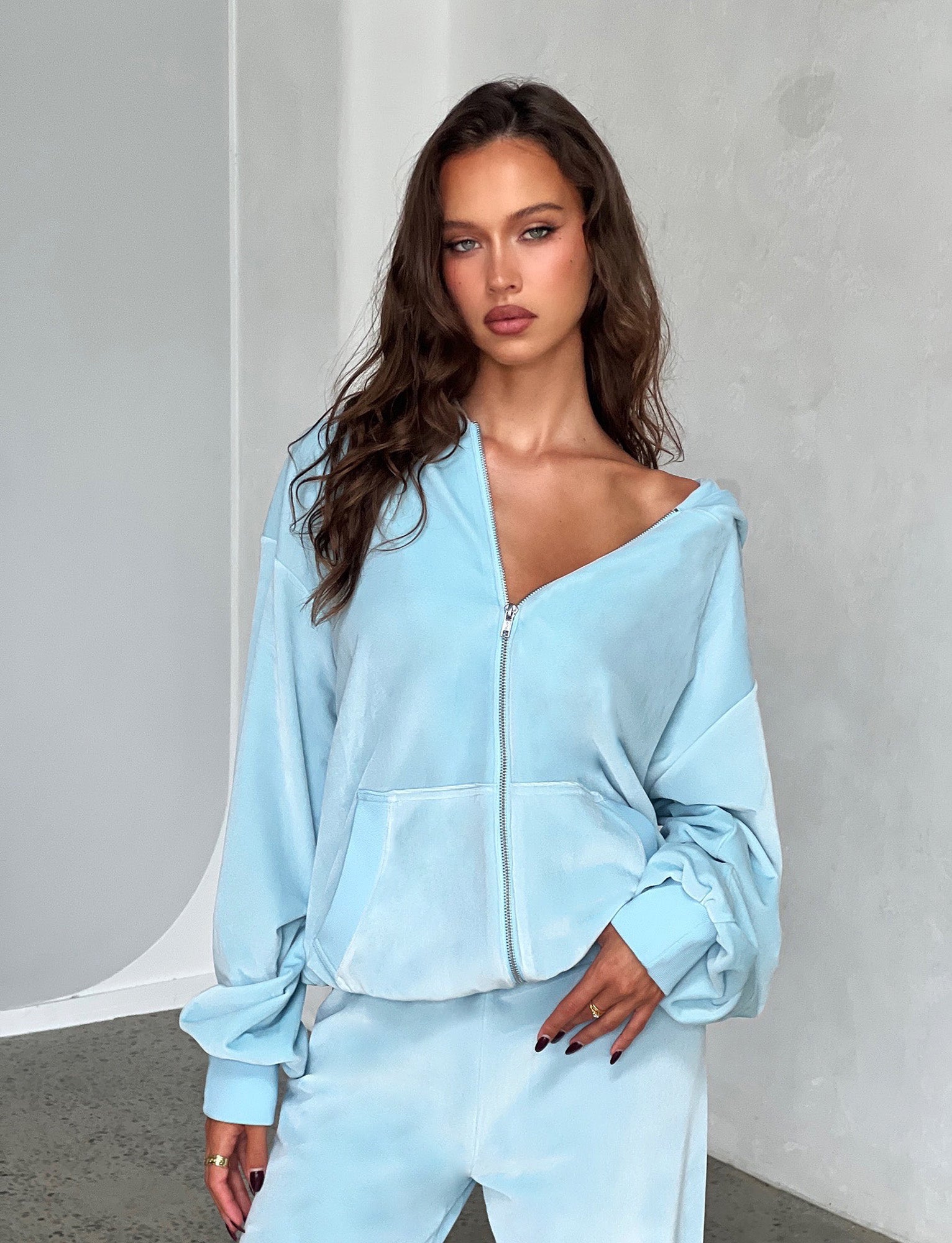 A woman with long, wavy brown hair poses against a neutral background. She is wearing a light blue TIGERMIST ERICKA HOODIE - BABY BLUE and matching pants. Her hand is resting on her hip, and she is looking directly at the camera with a neutral expression.