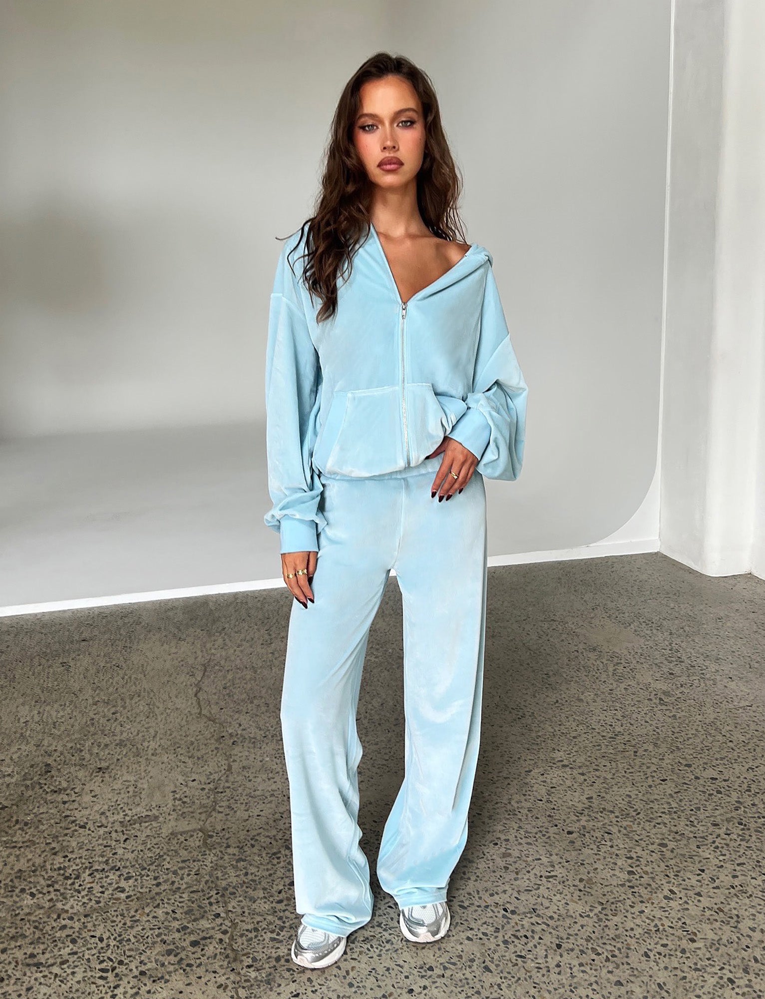 A woman stands indoors against a white backdrop, wearing the ERICKA HOODIE - BLUE: BABY BLUE from TIGERMIST, along with matching wide-leg pants. She has long, wavy brown hair and is posing with her hand in her pocket. She is also wearing white and gray sneakers.