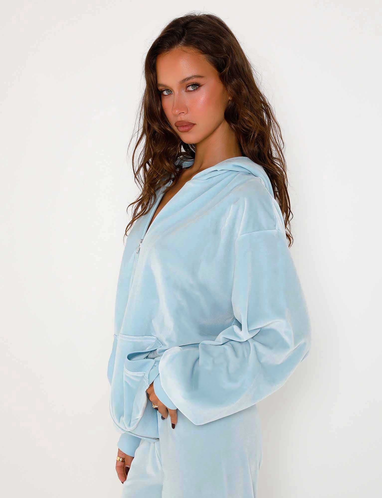 A woman with long, wavy brown hair stands against a plain white background, wearing the TIGERMIST ERICKA HOODIE in Baby Blue. She is looking confidently at the camera with her left hand resting on her waist.