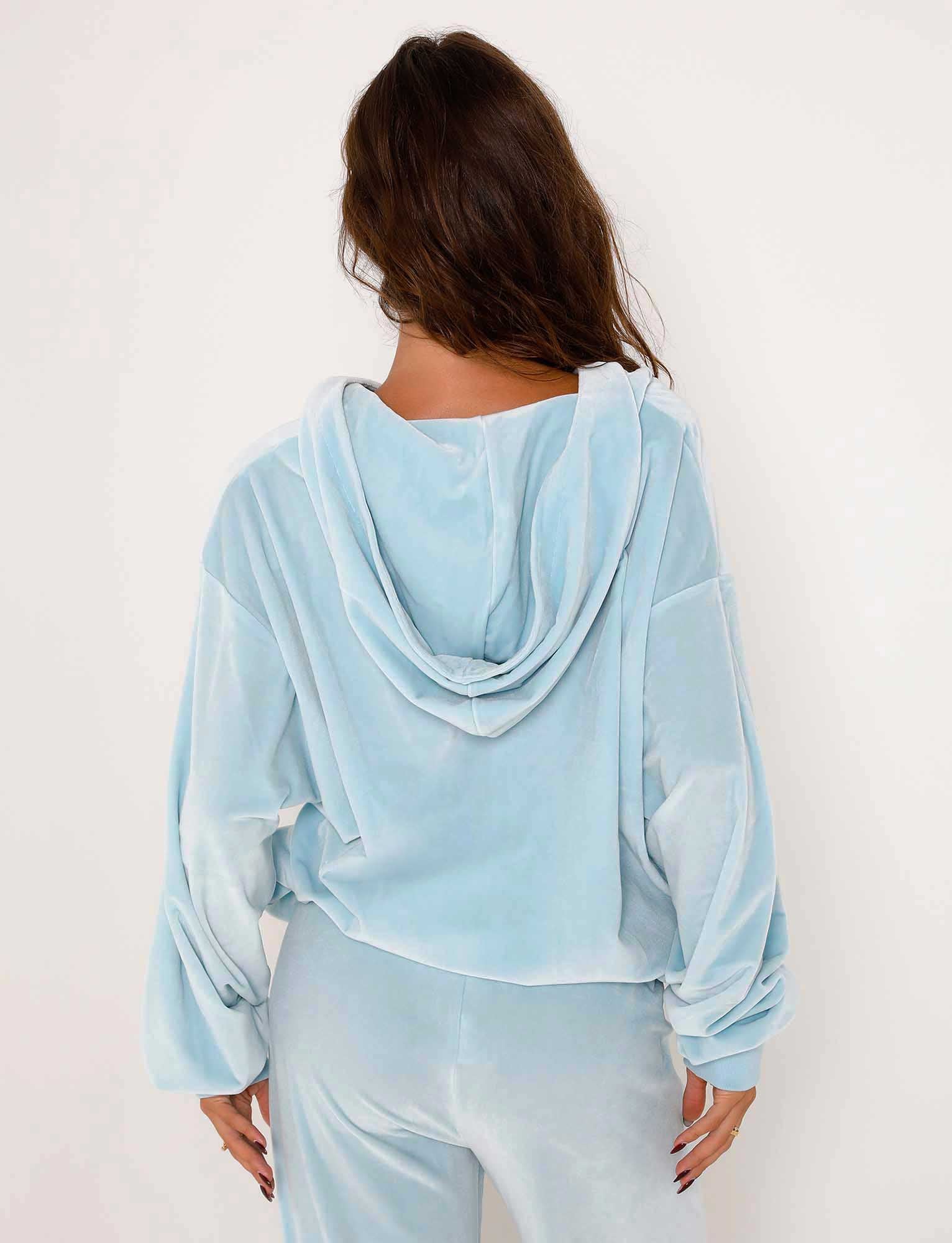 A woman with long brown hair is seen from behind, wearing the ERICKA HOODIE - BLUE : BABY BLUE by TIGERMIST. The loose-fitting top has a hood and appears soft and comfortable. She is standing against a plain white background.