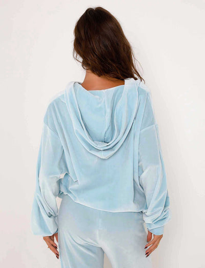 A woman with long brown hair is seen from behind, wearing the ERICKA HOODIE - BLUE : BABY BLUE by TIGERMIST. The loose-fitting top has a hood and appears soft and comfortable. She is standing against a plain white background.