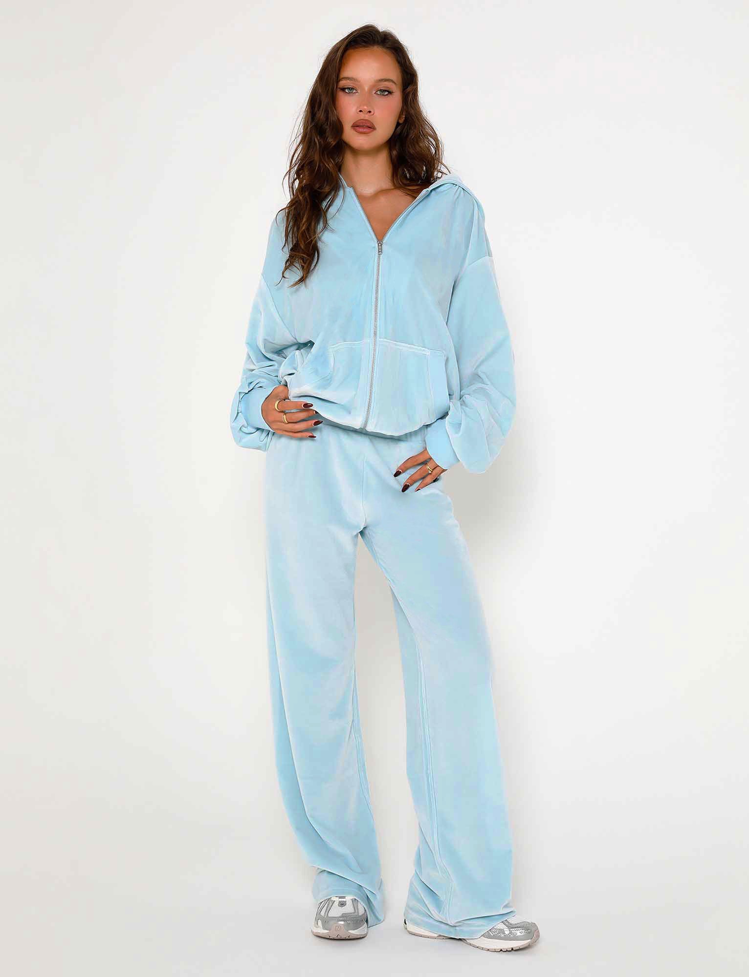 A person with long hair is wearing the TIGERMIST ERICKA HOODIE in baby blue along with matching baggy pants and silver sneakers. The tracksuit includes a zip-up hoodie with pockets. Standing against a plain white background, the person gazes towards the camera with one hand resting on their hip.
