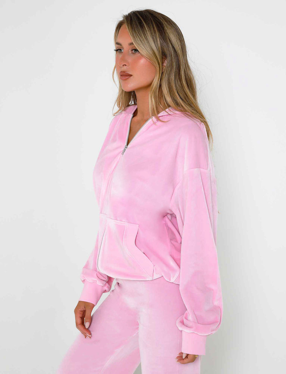 A woman with long blonde hair is standing in a three-quarter pose against a white background. She is wearing the Baby Pink ERICKA HOODIE and matching pants from TIGERMIST. The velour tracksuit includes a zipped hoodie with a front pocket, and she appears to be looking slightly to her right.
