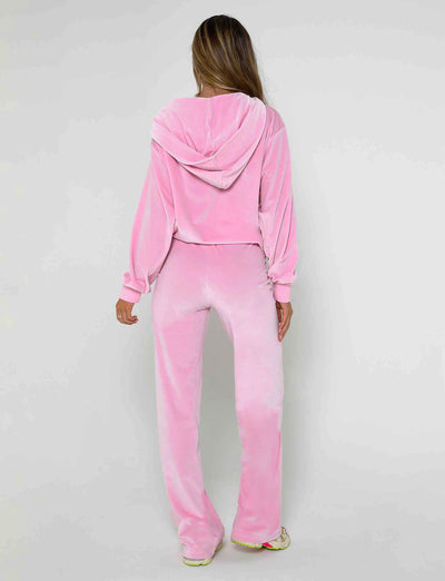 A person with long hair is standing and facing away from the camera, wearing a TIGERMIST pink velour tracksuit. The outfit includes a hooded top and the ERICKA PANT in baby pink. They are also wearing light-colored sneakers. The background is plain and light-colored.