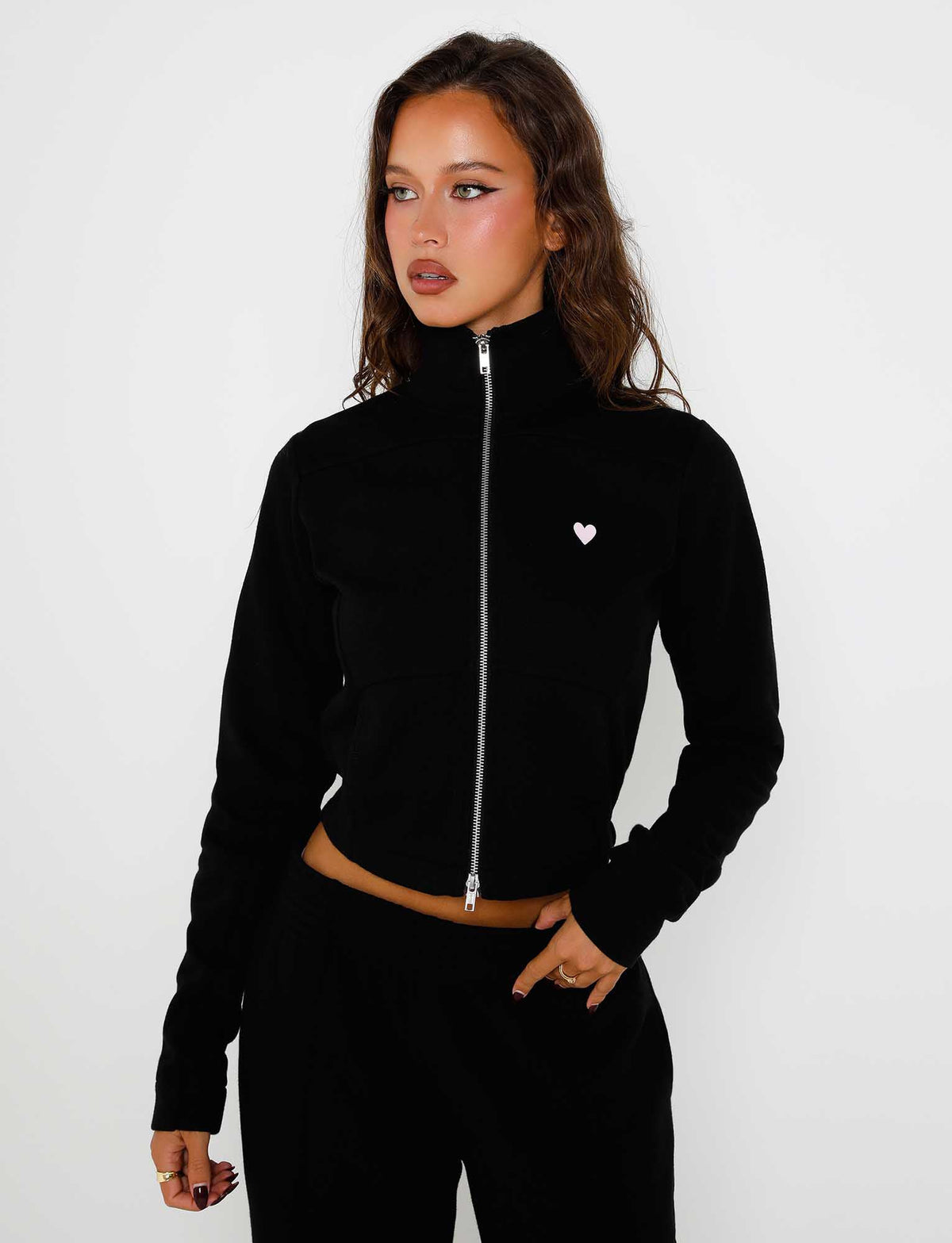 A woman with long dark hair is wearing the Tiger Mist VERDA jacket in black, featuring a small white heart emblem on the chest. She has a serious expression and is looking slightly to the side against a plain white background.