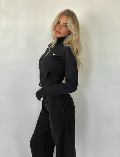 A woman with long blonde hair is posing against a plain white background. She is wearing the VERDA JACKET - BLACK and matching black pants from TIGERMIST. She is looking directly at the camera with a relaxed, confident expression.
