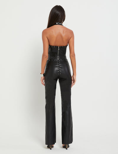 A person with long dark hair stands facing away from the camera, showcasing a stylish TIGERMIST BOSCO TOP in black PU and matching high-waisted pants with button details on the back. The background is plain white.