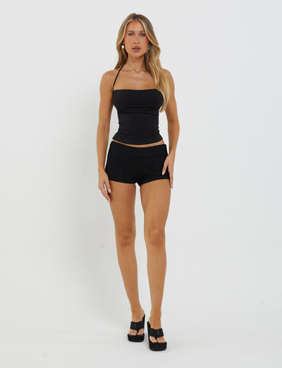 A woman with long blonde hair stands against a plain white background, dressed in a sleek black ZAIDA TOP by TIGERMIST and matching Tiger Mist shorts, complemented by black high-heeled sandals. She poses with her arms relaxed by her sides, looking straight at the camera.
