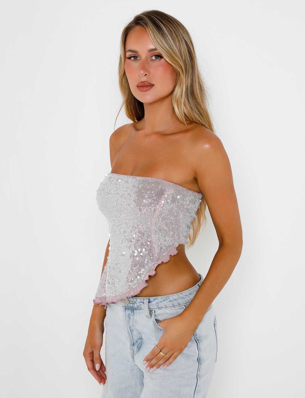 A woman stands against a white background, wearing the Opaline Top in Blush Sparkle from Tiger Mist, which features a strapless design and an asymmetrical hem. She pairs it with light-blue high-waisted jeans. Her long, wavy blonde hair and neutral expression complete the look.