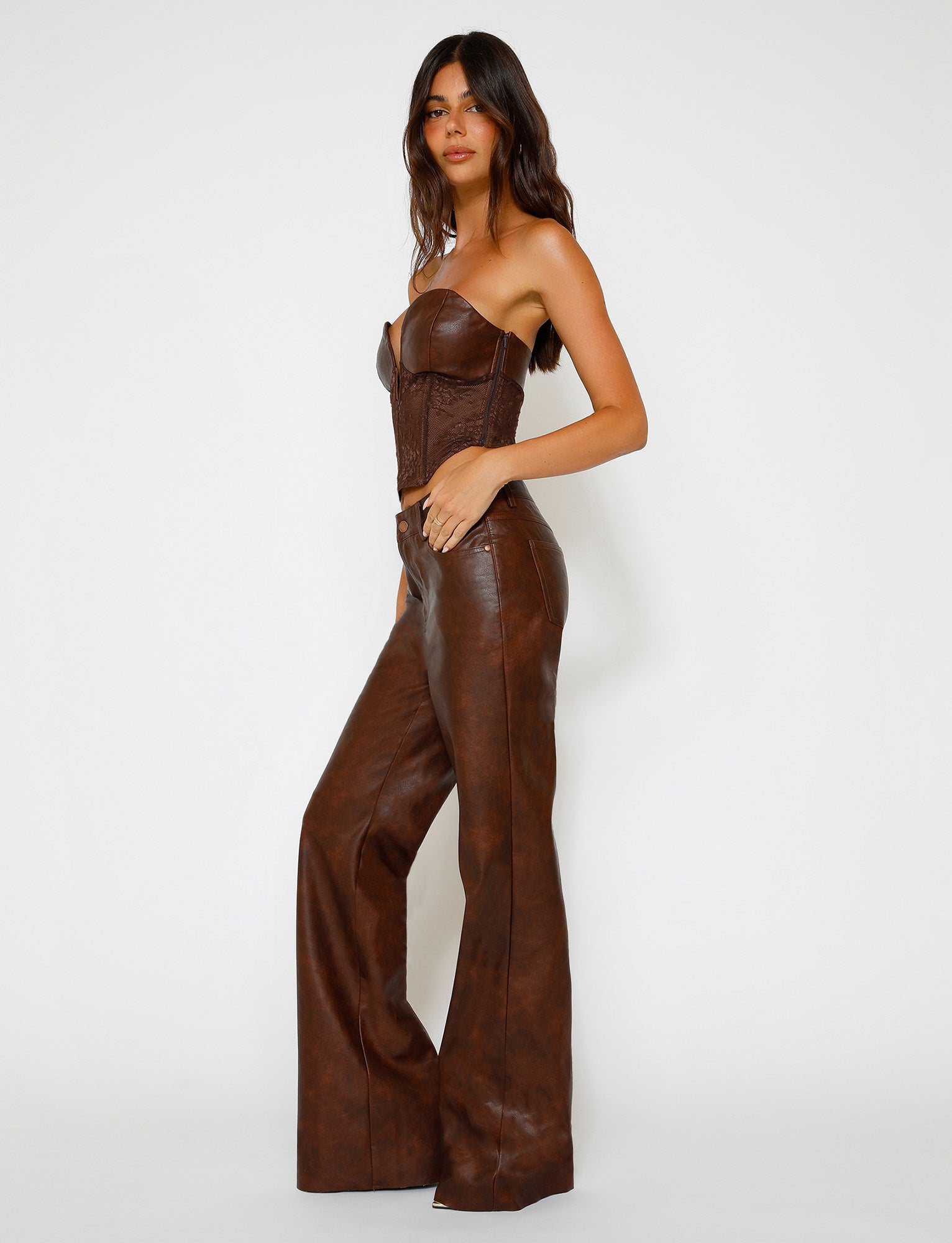 A woman stands against a plain white background, wearing a brown strapless corset top paired with high-waisted, wide-leg leather pants in Red Brown from TIGERMIST's WILDER PANT collection. She has long dark hair and is looking at the camera with a slight smile. Her right hand rests in her pants pocket.