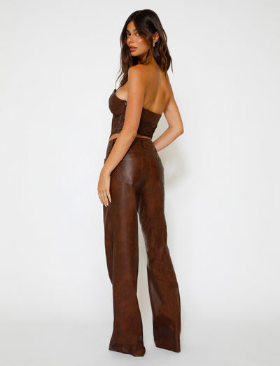 A woman with long, dark hair stands against a plain white background. She faces away from the camera, turning her head slightly to the left. Clad in a stylish TigerMist strapless brown top and matching high-waisted, wide-leg WILDER PANT in red-brown, she exudes effortless elegance.