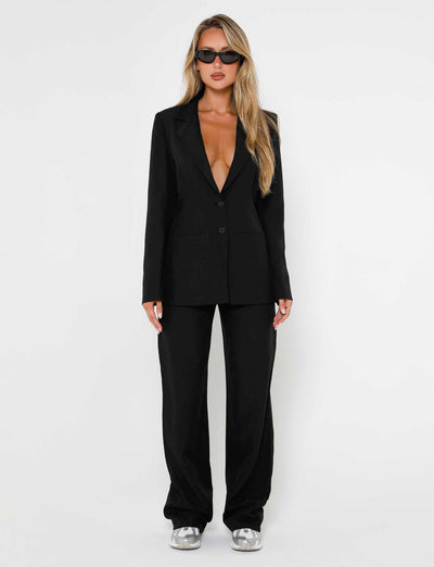 A woman stands against a white background wearing a sleek TIGERMIST MARJORIE JACKET - BLACK along with matching black pants. Her long blonde hair flows freely, and she sports dark sunglasses. Silver shoes complete her striking look.