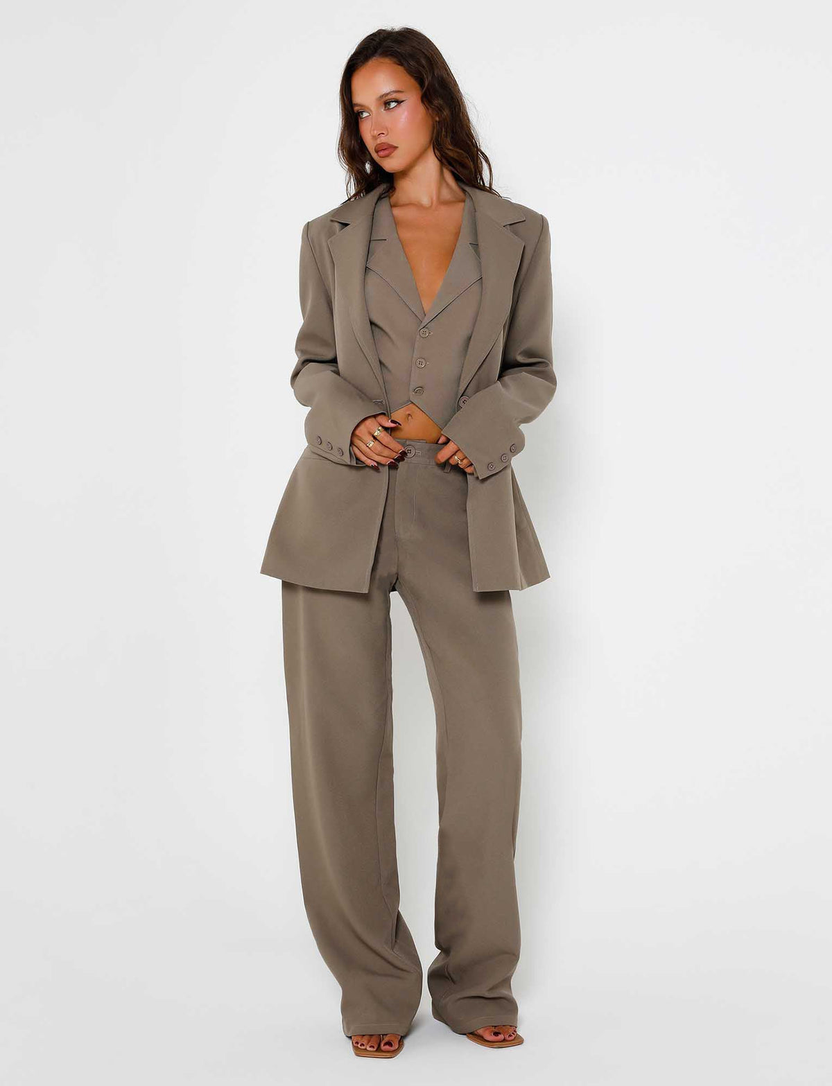 A woman stands against a plain white background, wearing the MARJORIE JACKET in moss green from TIGERMIST. She pairs the oversized blazer with a fitted vest and wide-leg trousers. Her long, wavy hair cascades down as she gazes off to the side, her hands casually tucked into the pockets of her blazer.