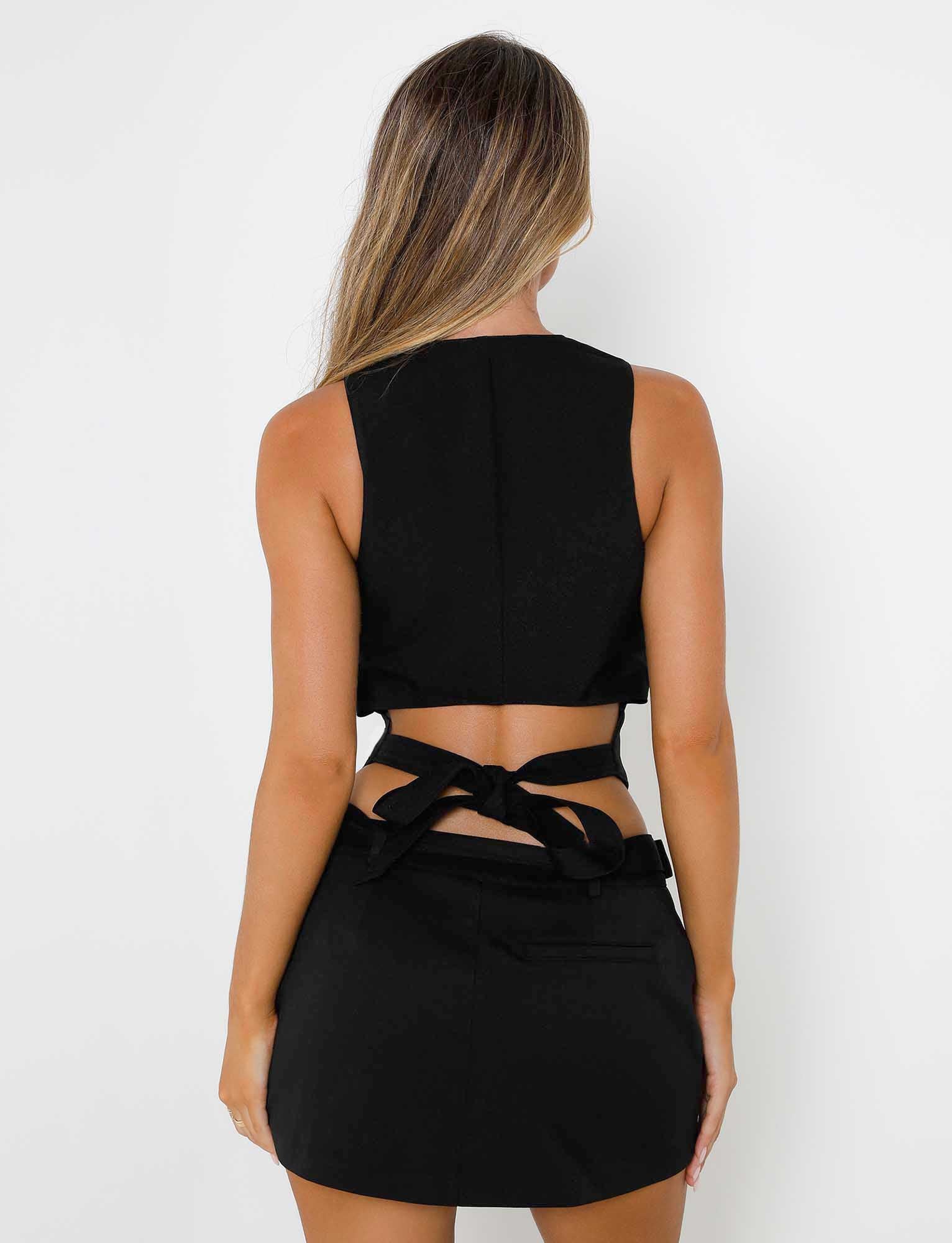 A woman with long hair is facing away from the camera, wearing the black MARJORIE VEST - BLACK and matching skirt from TIGERMIST. The back of her vest features criss-cross tie details at the waist. The background is white.