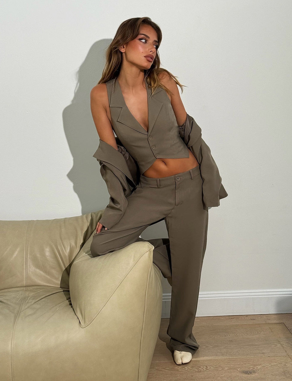 A woman poses stylishly in a moss green Marjorie Vest from Tigermist, featuring a sleeveless design and matching khaki trousers. She has her jacket draped off her shoulders and stands with one leg propped on a beige leather couch against a plain white wall.